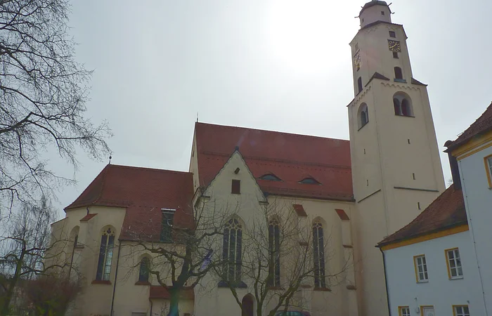 Kirchengebäude mit rotem Dach, einem Turm rechts im Bild mit Uhr und kahlen Bäumen mit grüner Wiese im Vordergrund bei bewölktem Himmel.
