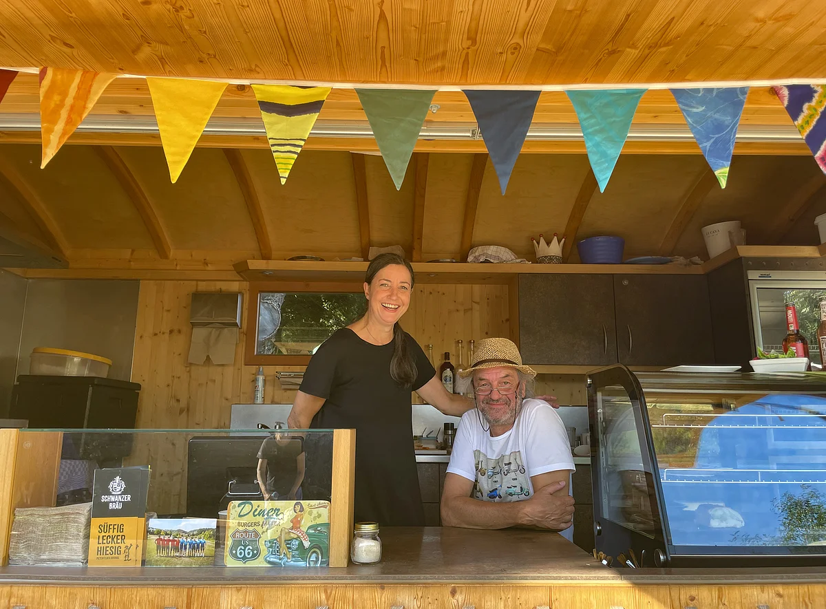 Zwei Personen in einem Holzverkaufsstand mit bunter Wimpelkette, eine Frau steht, ein Mann sitzt mit Strohhut.