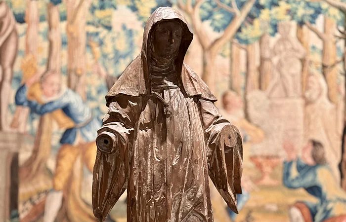 Vor einem Wandteppich mit vielen Baumverzierungen steht eine hölzerne Statue.