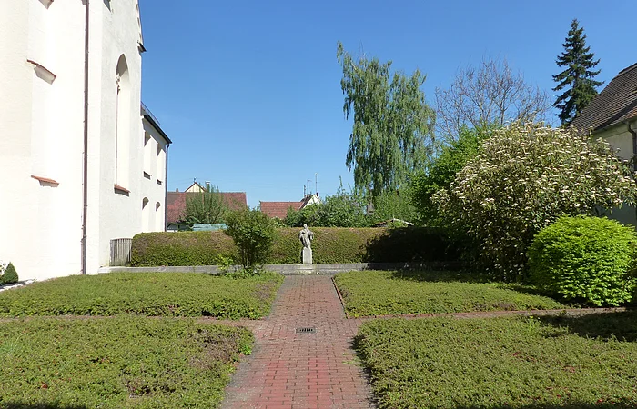 Ehemaliger Klostergarten in Monheim mit gepflastertem Weg in der Mitte, niedrigen Hecken, Büschen und Bäumen. Am Ende des Weges eine Statue vor einer Hecke unter blauem Himmel. Links im Bild ein weißes Gebäude.