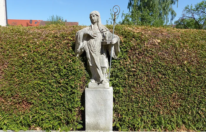 Steinerne Statue der Heiligen Walburga mit Stab vor einer hohen Hecke und blauem Himmel.