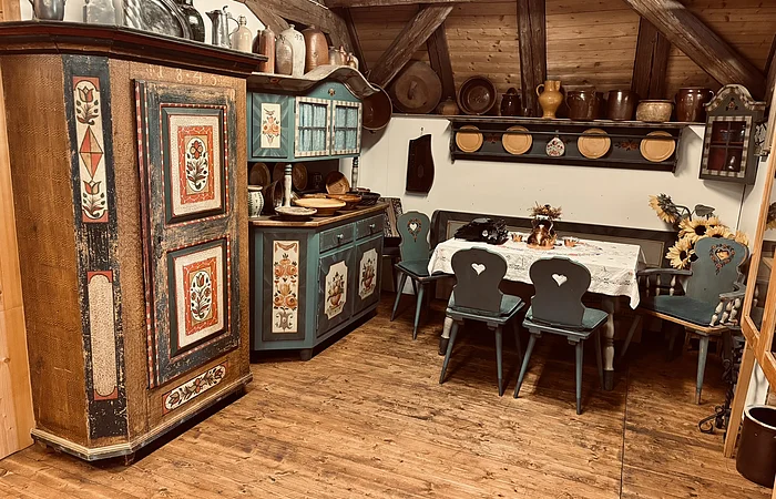 Links sind zwei alte Bauernschränke mit bunten Verzierungen zu sehen. Rechts ein Tisch mit Stühlen
