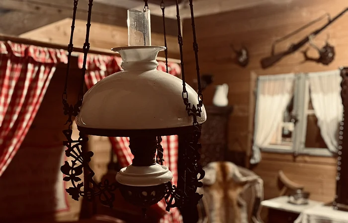 In der Mitte des Bildes hängt eine Öllampe von der Decke über einem Tisch. Im Hintergrund ist eine Fensterbank mit roten Vorhängen zu sehen.