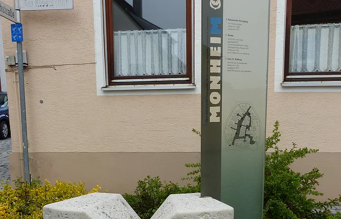 Steinerner grauer Buchstabe M vor Infotafel aus Glas mit Erklärtext und einem Stadtplan. Auf der Infotafel vertikal der Text „Monheim“. Im Hintergrund ein Haus in beige mit einer Hecke und Straßenschildern.
