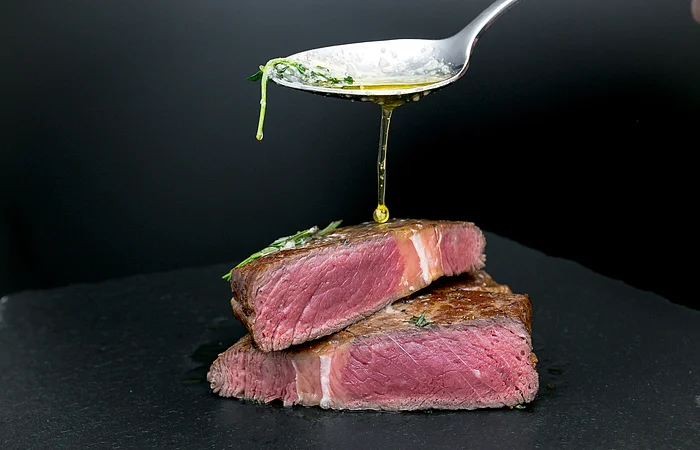 Ein Löffel gießt Öl über zwei aufeinander gestapelte, halbierte Rindersteaks auf einem schwarzen Hintergrund.