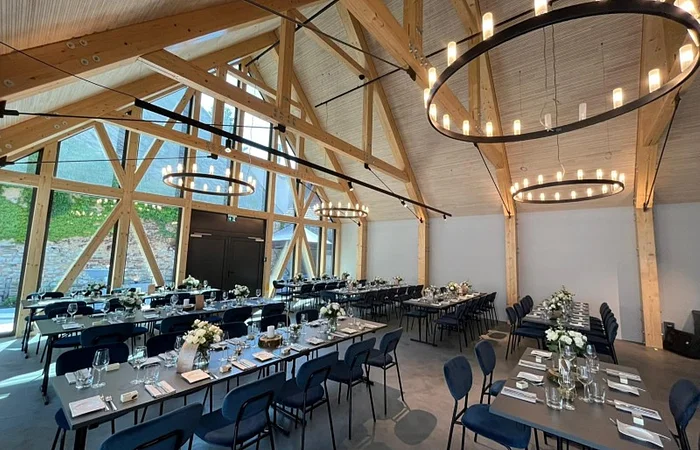 Großer Veranstaltungsraum einer Eventlocation mit offenem Dachstuhl und sichtbaren hellen Holzbalken oben an der Decke. In Reihen aufgestellte schwarze Tische mit denkelblauen Stühlen. Tische sind gedeckt mit Gläsern, Servietten, Besteck und Blumen in Vasen. Links im Bild große Glasfront ebenfalls mit hellen Balken. An der Decke große, runde, moderne Leuchten angebracht.