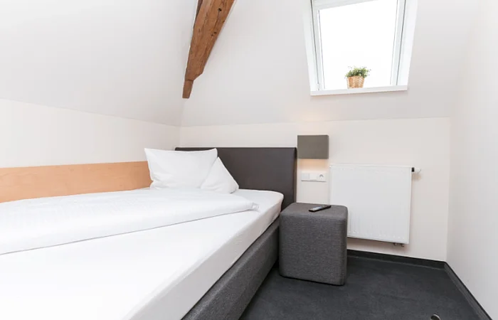 Einzelzimmer in einem Hotel mit einem Einzelbett links an der weißen Wand, einem grauem Hocker rechts daneben, auf dem eine  Fernbedienung liegt und einer grauen Leuchte oberhalb an der Wand. Dachschräge mit Holzbalken, rechts davon und kleines Fenster mit Pflanze auf dem Fensterbrett. Unter dem Fester eine kleine weiße Heizung an der Wand angebracht.