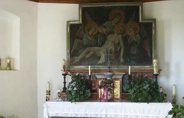 Innenansicht einer Kapelle: Altar mit weißem Tuch bedeckt, umgeben von verschiedenen Kerzen, Blumen und religiösem Gemälde oberhalb an der Wand, in einem Raum mit Holzbalkendecke.