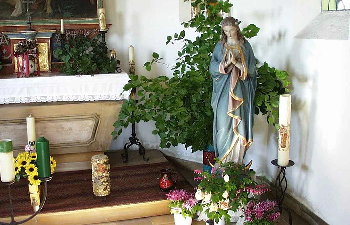 Innenansicht einer kleinen Kapelle mit der Statue einer betenden Frau mit blauem Mantel in einer Ecke umgeben von Kerzen, Pflanzen und Blumen in einem weißen Raum. Links im Bild ein geschmückter Altar.