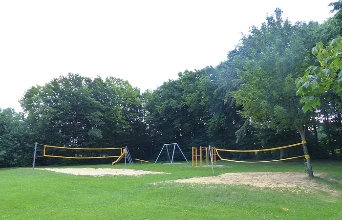 Spielplatz mit zwei gelben Volleyballnetzen, einer Rutsche, einer Schaukel und Klettergerüst vor Baumreihe auf Grasfläche, bei Tageslicht.