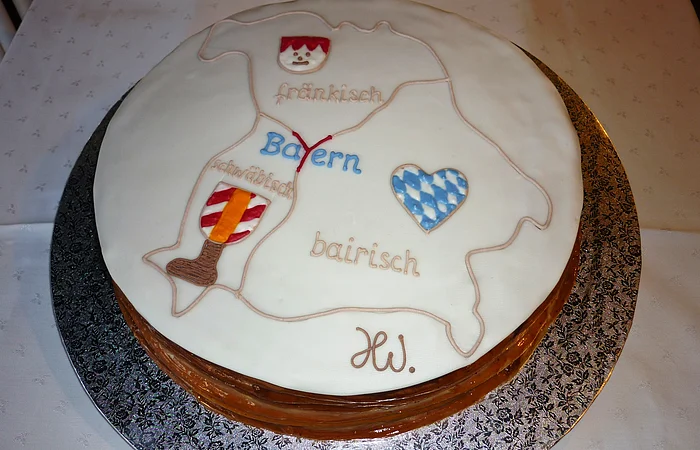 Runde Torte auf einer silbernen Unterlage seht auf einem Tisch mit weißem Tischtuch. Auf der Torte weiße Glasur und eine Karte von Bayern mit Dialektbezeichnungen und den dazugehörigen Symbolen darauf.