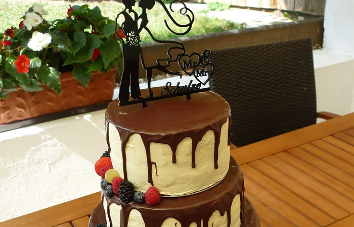 Vierstöckige Hochzeitstorte mit runterlaufender Schokoladenglasur und weißer Creme, verschiedenen Beeren auf drei Stockwerken und einem Brautpaar-Topper mit Schriftzug „Mr & Mrs“ oben drauf. Im Hintergrund ein Garten mit bepflanzten Blumentöpfen und grüner Wiese.