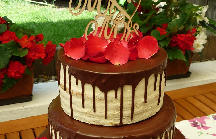 Dreistöckige Hochzeitstorte mit runterlaufender Schokoladenglasur auf allen drei Stockwerken, drei roten Rosen unten und dem Schriftzug „Mrs & Mrs“ aus Holz obenauf. Im Hintergrund ein Garten mit bepflanzten Blumentöpfen und grüner Wiese.