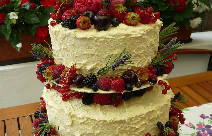 Dreistöckige Torte mit weißer Creme, dekoriert mit Beeren, Kirschen, Lavendel und Rosmarinzweigen steht auf einem Tisch. Im Hintergrund ein Garten mit bepflanzten Blumentöpfen und grüner Wiese.