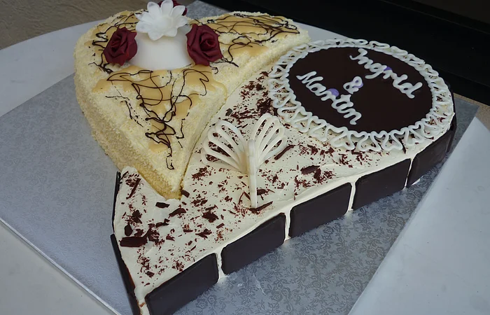 Herzförmige Hochzeitstorte mit weißer und dunkler Schokolade, beschriftet mit „Ingrid & Martin“ und dekoriert mit zwei roten Rosen und verschiedener Schokoladen-Dekoration, steht auf einer silbernen Unterlage auf einem Tisch.