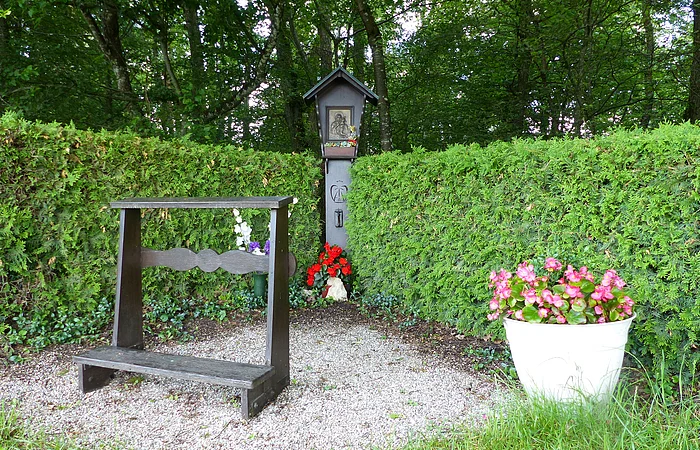 Begrenzter Gebetsplatz mit Holzkneeler auf Schottersteinen, Blumen und kleinem Bildstock vor grüner Hecke im Wald.