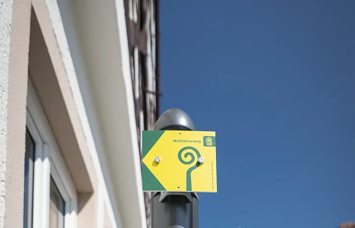Drei leuchtend gelbe Schilder am grauen Pfosten in einer Altstadt bei blauem Himmel. Zwei Schilder zu Wanderwegen mit der Nummern 5 und 6 und ein Pilgerweg-Symbol für den Wallfahrerweg, mit dem dazugehörigen spiralförmigen Symbol.