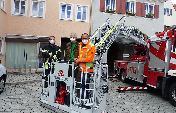 Drei Personen mit Schutzmasken stehen auf einer Feuerwehr-Hubarbeitsbühne vor einem roten Feuerwehrfahrzeug mit ausgefahrener Leiter. Im Hintergrund zwei farbige Gebäude und gepflasterter Platz vorne.