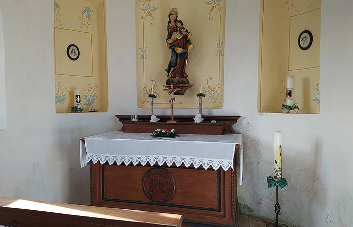 Kleiner Kirchenaltar einer Kapelle mit Marienstatue, Kerzen und Blumenschmuck vor weiß-gelb gestrichener Wand mit Ausfräsungen in der Wand mit gemalten Verzierungen darin. Im Vordergrund eine Gebetsbank aus Holz.