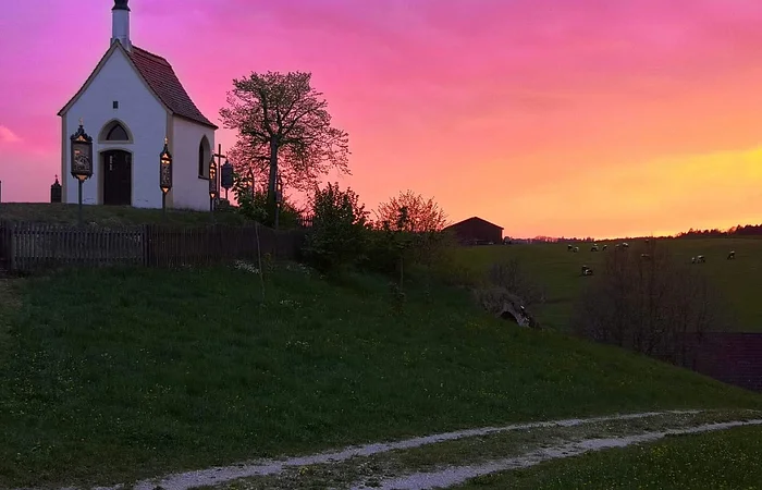 Links im Bild eine kleine Kapelle auf einem Hügel mit einem Weg unten am Hügel, umgeben von Bäumen und Weide. Sonnenuntergang mit pink-orangefarbenem Himmel im Hintergrund.
