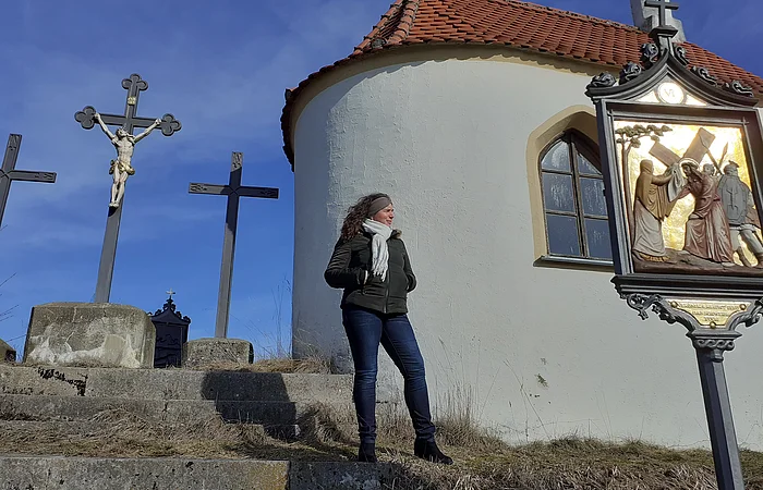 Frau steht auf einer Steintreppe neben einer Kapelle mit drei Kreuzen im Hintergrund und einem religiösem Bild auf einer Tafel, bei blauem Himmel.