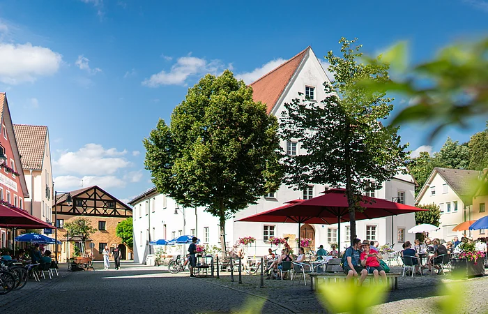 Platz mit historischen Häusern, Cafés und großen Sonnenschirmen, im Vordergrund ein Paar mit Smartphone auf einer Bank