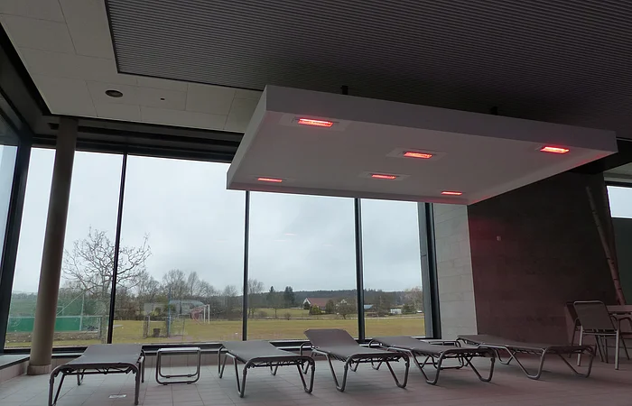 Innenraum eines Hallenbades mit Liegestühlen vor großen Fenstern und einer Deckenleuchte mit roten Lichtern.