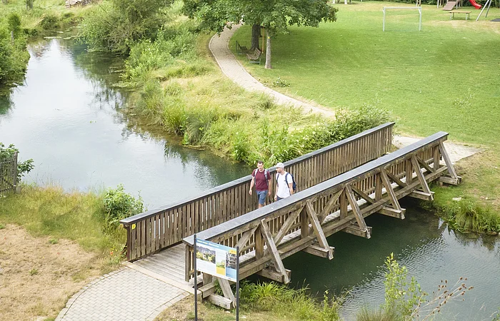 Drohnenaufnahme von Möhren. Im Vordergrund sind 2 Wanderer auf einer Holzbrücke über den Möhrenbach zu sehen. Im Hintergrund thront Schloss Möhren über dem Dorf.