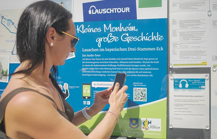 Frau mit Smartphone steht vor Infotafel zur Lauschtour in Monheim, Bayern, mit dem Titel "Kleines Monheim, große Geschichte" bei Sonnenschein. Frau hält ihr Smartphone an einen QR-Code.