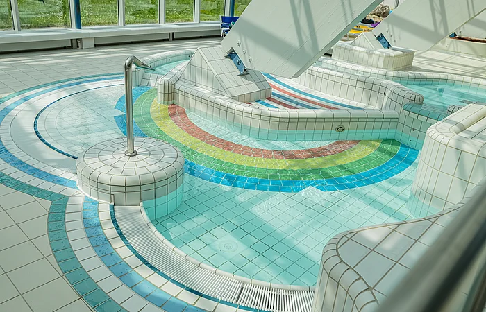 Innenbereich eines Schwimmbads mit kleinem Becken und bunten Fliesen in Regenbogenfarben am Boden.