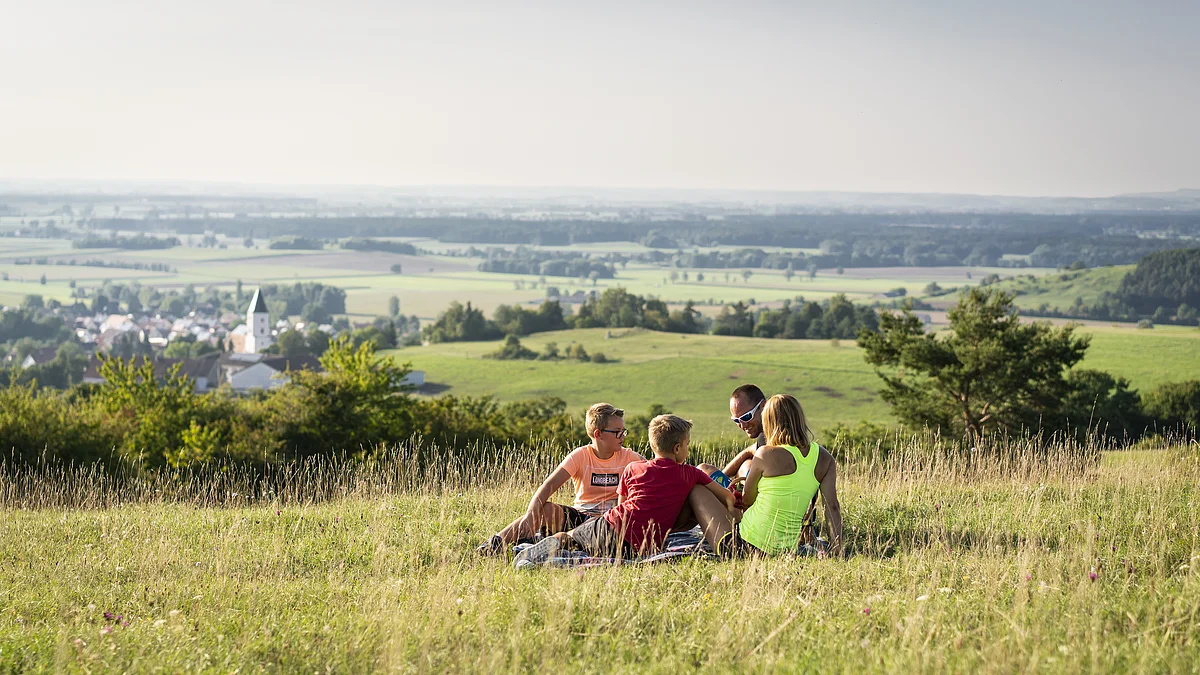 Vier Personen sitzen auf einer Wiese mit Blick auf ein Dorf und eine weite Landschaft im Hintergrund.