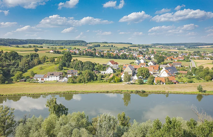 Dorf mit Häusern, Feldern und Bäumen an einem Fluss unter blauem Himmel mit Wolken