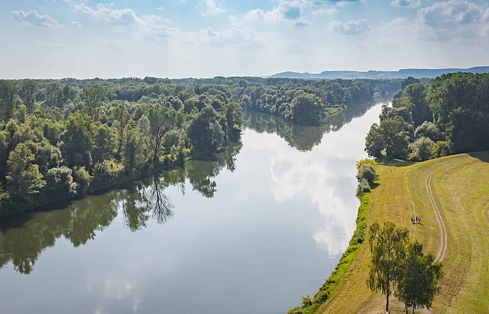 Fluss mit bewaldetem Ufer links und Wiese mit Wanderern rechts unter bewölktem Himmel