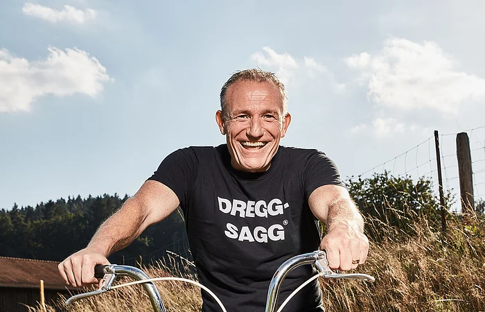 Mann mit schwarzem T-Shirt mit der weißen Aufschrift "DREGG-SAGG" und Jeans sitzt lachend auf orangefarbenem Fahrrad auf Feldweg bei Sonnenschein