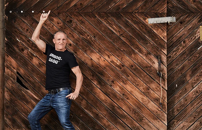Mann in schwarzem T-Shirt mit Aufschrift „DRIGG-SAGG“ und dunkelblauer Jeans posiert in die Kamera blickend vor Holztür mit diagonalem Muster.
