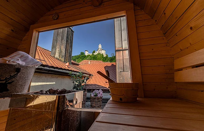 Blick aus einer Holzsauna auf ein geöffnetes Fenster mit Dachziegeln und einer Burg auf einem Hügel im Hintergrund.