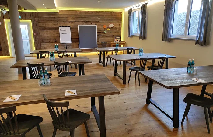 Konferenzraum mit Holztischen, Stühlen, Wasserflaschen und Flipchart vor Holzpaneelwand und Fenstern mit Vorhängen.