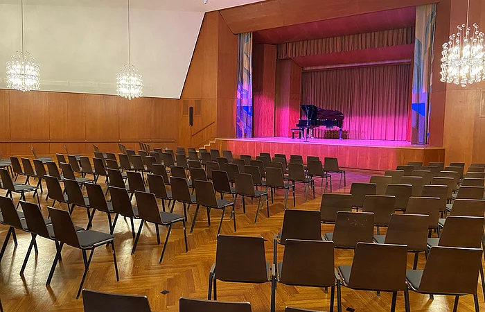 Leerer Konzertsaal mit Reihen von Stühlen und einem Flügel auf der Bühne mit roter Beleuchtung.