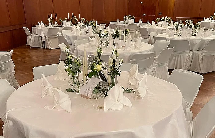 Rund gedeckter Tisch mit weißen Hussen, Servietten, Kerzen und Blumen in einem Saal mit Holzwänden.