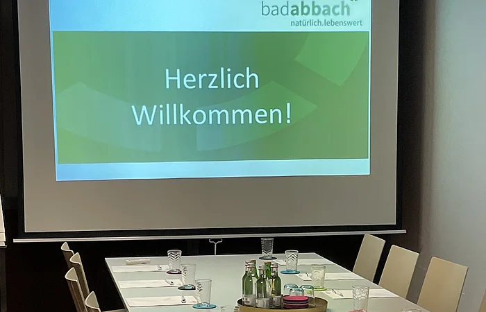 Konferenzraum mit langem Tisch, Stühlen, Gläsern, Flaschen und Projektionsbild mit Text „Herzlich Willkommen!“