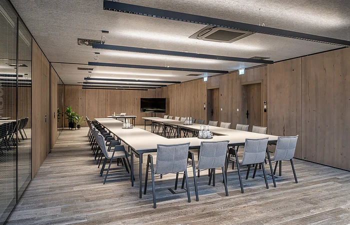 Konferenzraum mit U-förmigen Tischen, grauen Stühlen, Holzwänden und Deckenleuchten.