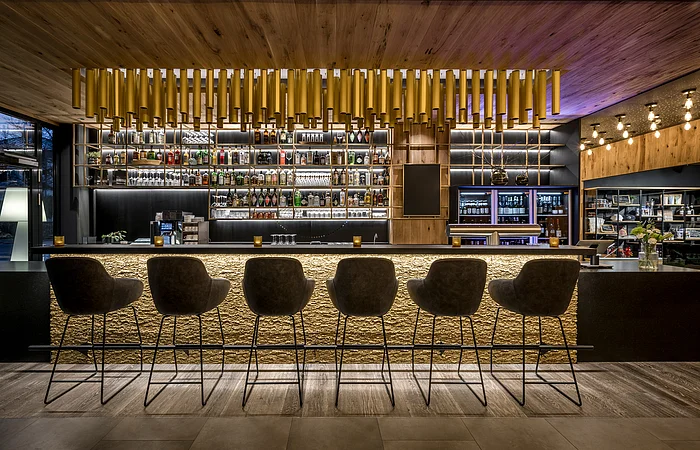 Bar mit sechs hohen Stühlen, beleuchteter Tresen und Regal mit Flaschen und Gläsern im modernen Innenraum.