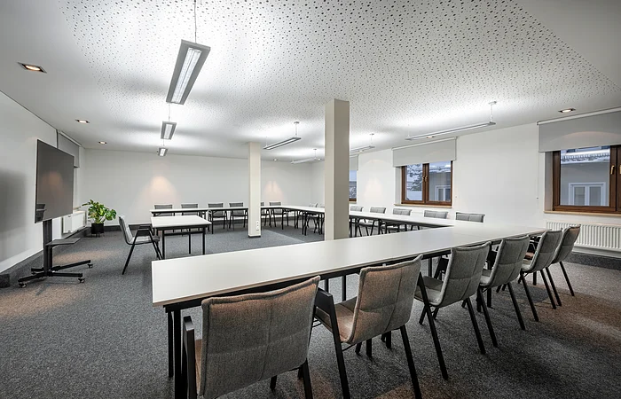 Großer heller Konferenzraum mit U-förmigen Tischen, grauen Stühlen, Fenstern und Deckenleuchten.