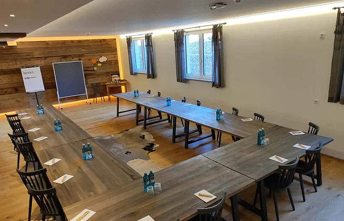 Konferenzraum mit U-förmigen Holztischen, Stühlen, Wasserflaschen und Schreibblöcken bei Tageslicht.