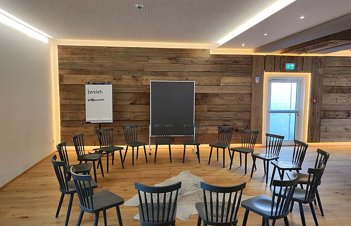 Konferenzraum mit Holzfußboden, Stühlen im Kreis, Flipchart mit „herzlich willkommen“ und Holzwand mit Tür und Tafel.