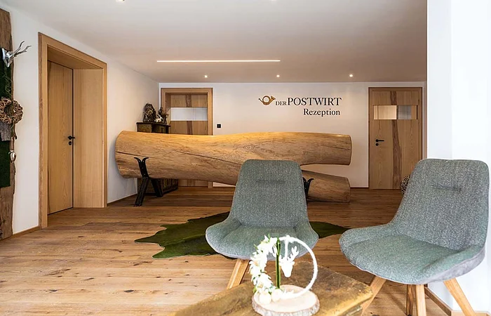 Empfangsbereich mit rustikalem Holztresen, zwei grauen Stühlen und Holzboden, Wand mit Schriftzug „Der Postwirt Rezeption“