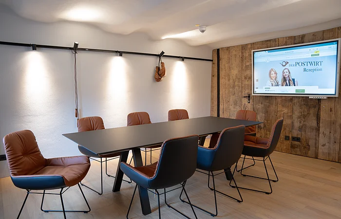 Konferenzraum mit sechs braunen Stühlen um schwarzen Tisch, Holzwand und Bildschirm an weißer Wand.