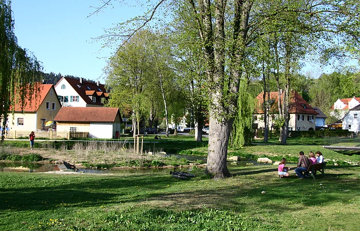 Grüne Parkwiese mit Bäumen, Bach, Fahrrad und Menschen auf Bank vor Häusern unter blauem Himmel.