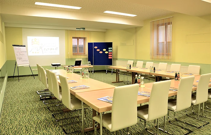 Tagungsraum mit U-förmiger Tischanordnung, weißen Stühlen, Flipchart und Projektor mit Leinwand.