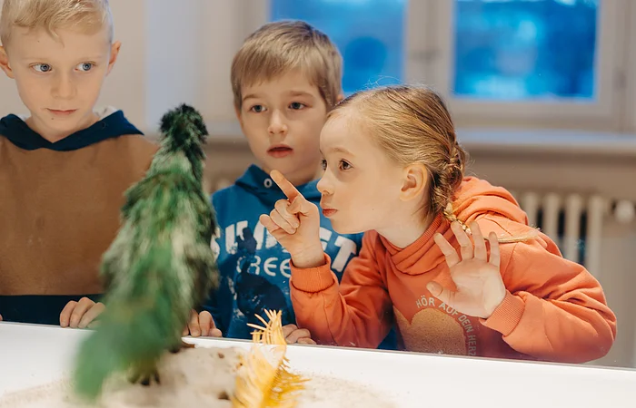 Drei Kinder beobachten aufmerksam ein grünes, federartiges Objekt hinter einer Glasvitrine.